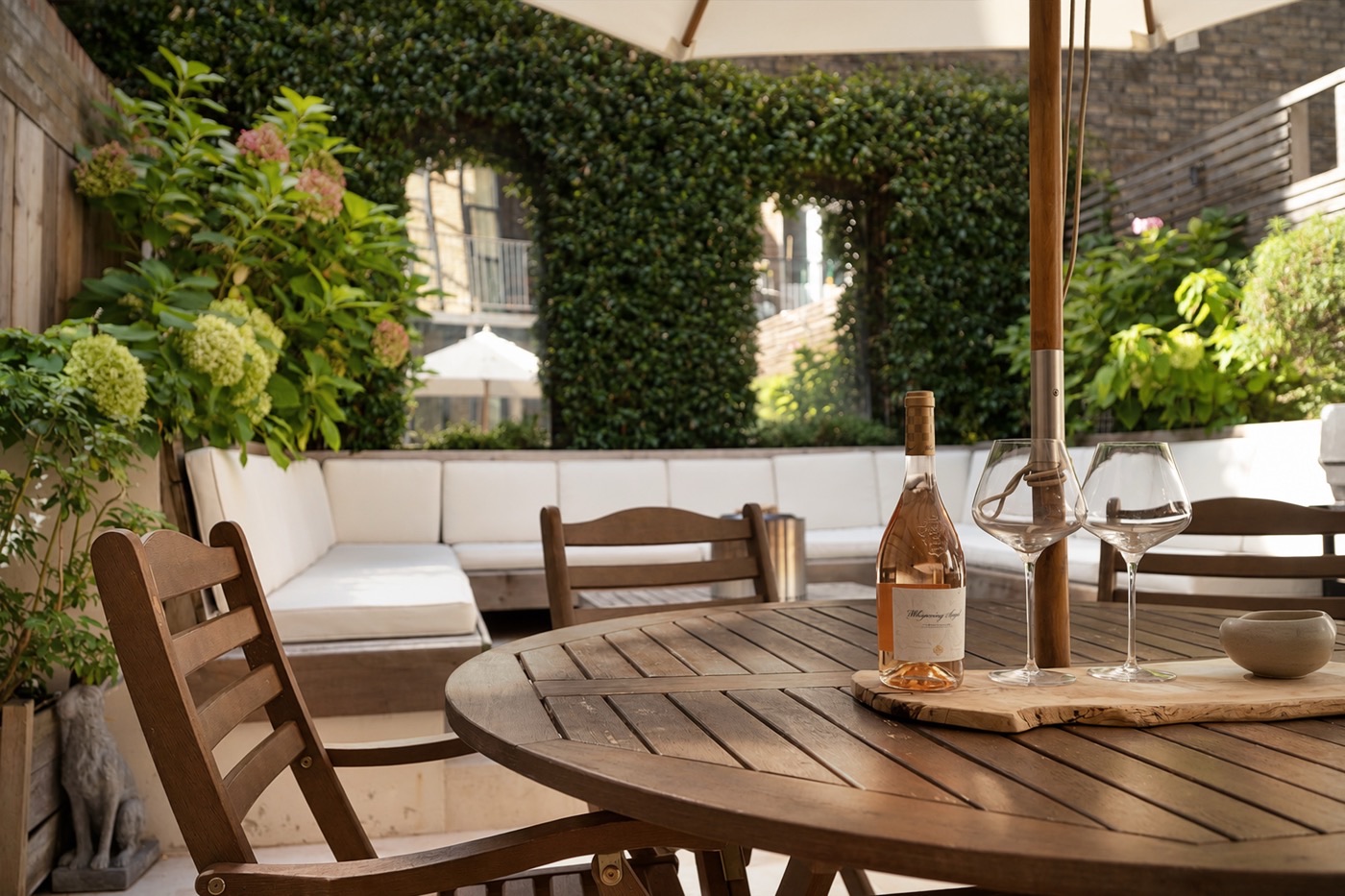 A wooden garden table set with a bottle of rosé and two glasses under a parasol, with ivy-covered walls and built-in seating behind, photographed by Fulham Performance